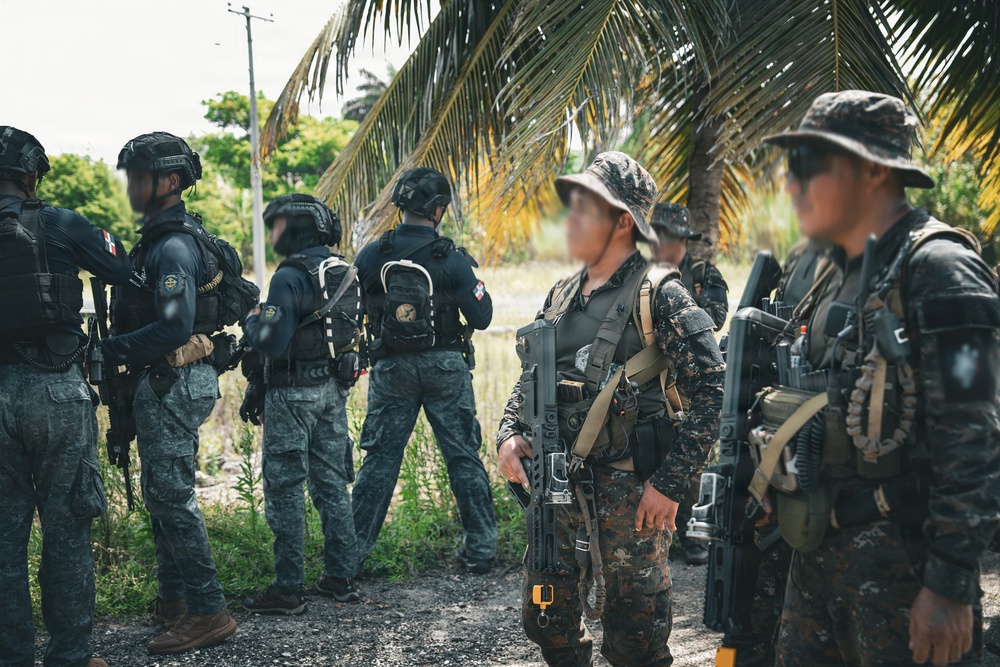 A group of armed security or military personnel are assembled in an outdoor tropical setting near palm trees. The individuals wear mixed uniforms including dark tactical gear and camouflage patterns, and are equipped with firearms and tactical equipment. Faces have been deliberately blurred, suggesting operational security or privacy considerations.