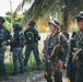 A group of armed security or military personnel are assembled in an outdoor tropical setting near palm trees. The individuals wear mixed uniforms including dark tactical gear and camouflage patterns, and are equipped with firearms and tactical equipment. Faces have been deliberately blurred, suggesting operational security or privacy considerations.