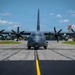 A large gray military turboprop aircraft, consistent with a C-130J Super Hercules variant, is taxiing or stationary on a runway/taxiway facing directly toward the camera. The aircraft features four turboprop engines with six-bladed propellers in motion, painted in a flat gray military finish. The setting appears to be a military airfield with green grass margins, runway markings, and support infrastructure visible in the background.