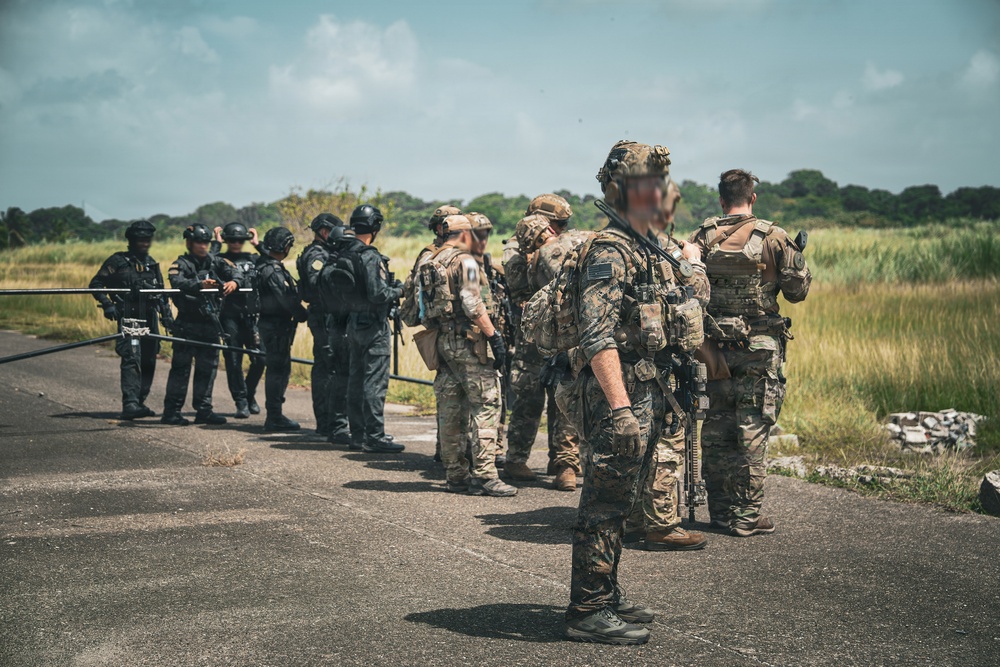A group of military and law enforcement personnel are gathered on what appears to be a tarmac or paved surface in a tropical or subtropical environment. Two distinct groups are visible: personnel in tan/multicam camouflage uniforms with tactical gear, and others in dark/black tactical uniforms. The scene appears to depict a joint briefing or coordination meeting between allied forces.