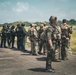 A group of military and law enforcement personnel are gathered on what appears to be a tarmac or paved surface in a tropical or subtropical environment. Two distinct groups are visible: personnel in tan/multicam camouflage uniforms with tactical gear, and others in dark/black tactical uniforms. The scene appears to depict a joint briefing or coordination meeting between allied forces.