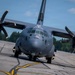 A large military turboprop transport aircraft, identifiable as a C-130J Super Hercules, is taxiing on a runway or taxiway. The aircraft is painted in a dark gray military finish and is photographed from a low front-facing angle, emphasizing its four six-bladed propellers. The setting appears to be a military airfield surrounded by dense coniferous trees under a partly cloudy sky.