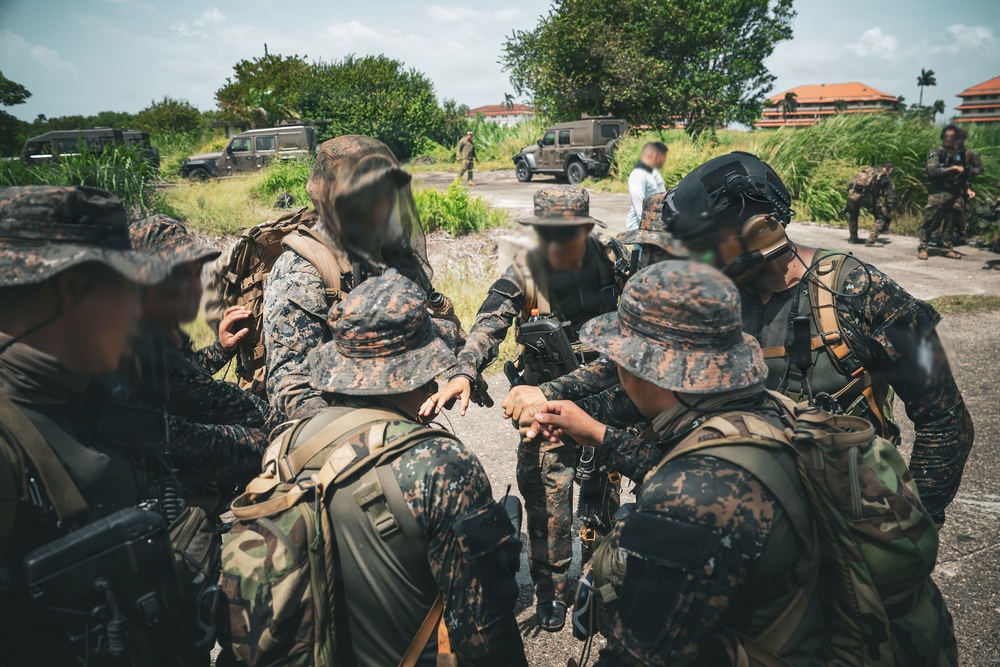 A group of military personnel in camouflage uniforms are gathered in a huddle, appearing to conduct a briefing or coordination meeting outdoors. The setting appears to be a tropical or subtropical environment based on the lush vegetation and architecture visible in the background. Multiple military vehicles and additional personnel are visible in the background.