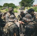 A group of military personnel in camouflage uniforms are gathered in a huddle, appearing to conduct a briefing or coordination meeting outdoors. The setting appears to be a tropical or subtropical environment based on the lush vegetation and architecture visible in the background. Multiple military vehicles and additional personnel are visible in the background.