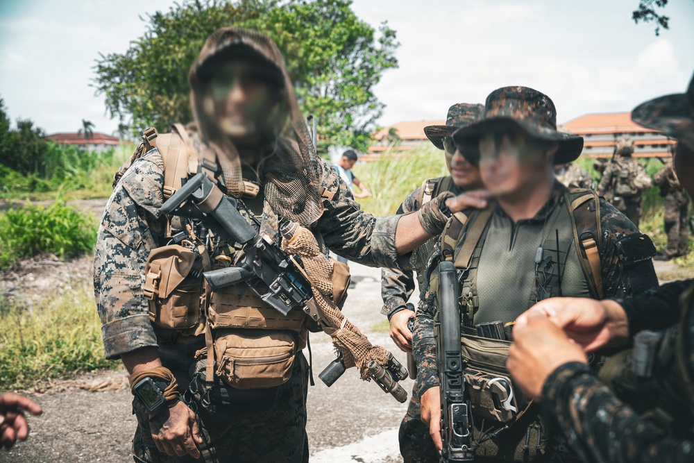 The image shows multiple armed personnel in military/tactical gear standing in an outdoor area with vegetation and buildings in the background. The individuals are wearing camouflage uniforms and carrying weapons and tactical equipment. Faces have been digitally obscured/blurred, suggesting privacy or operational security considerations.