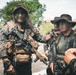 The image shows multiple armed personnel in military/tactical gear standing in an outdoor area with vegetation and buildings in the background. The individuals are wearing camouflage uniforms and carrying weapons and tactical equipment. Faces have been digitally obscured/blurred, suggesting privacy or operational security considerations.