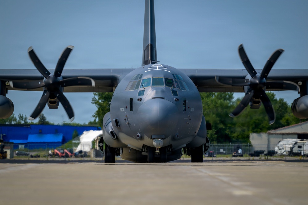A large military turboprop aircraft, identified by the number '5870' on its fuselage, is photographed head-on while on a tarmac or ramp area. The aircraft appears to be a C-130J Super Hercules or similar variant, featuring four-blade propellers and a gray low-visibility paint scheme. The setting appears to be a military or civilian airfield with trees, buildings, and equipment visible in the background.