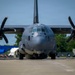 A large military turboprop aircraft, identified by the number '5870' on its fuselage, is photographed head-on while on a tarmac or ramp area. The aircraft appears to be a C-130J Super Hercules or similar variant, featuring four-blade propellers and a gray low-visibility paint scheme. The setting appears to be a military or civilian airfield with trees, buildings, and equipment visible in the background.