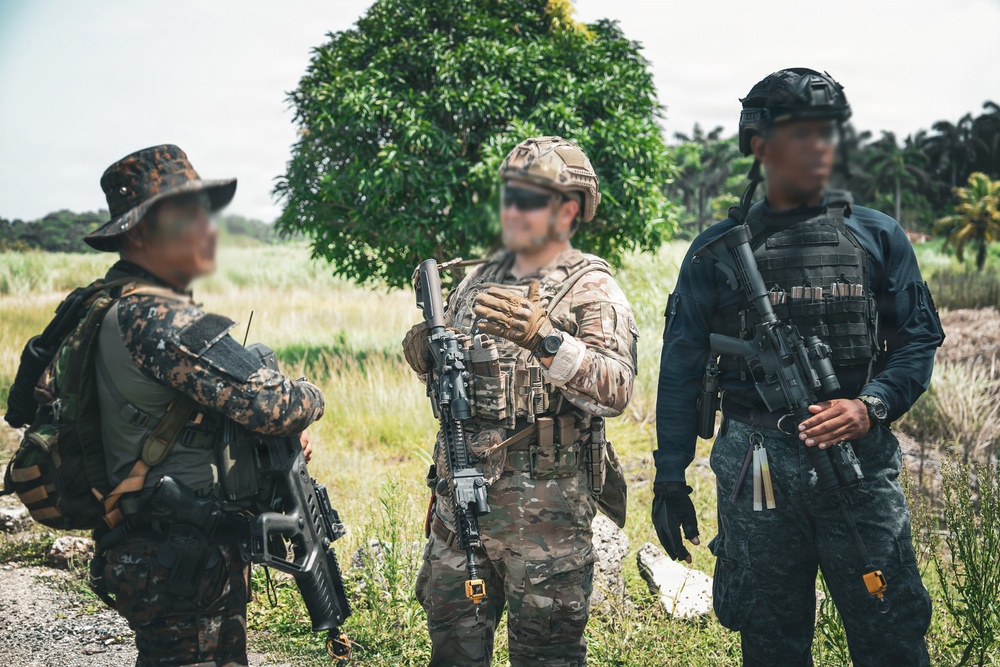 Three armed military or law enforcement personnel are standing in an outdoor field environment, appearing to be in discussion or coordination. They are wearing different uniforms and equipment, suggesting a multinational or joint-force operation. The faces of all individuals have been digitally blurred, indicating privacy or operational security measures.