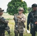 Three armed military or law enforcement personnel are standing in an outdoor field environment, appearing to be in discussion or coordination. They are wearing different uniforms and equipment, suggesting a multinational or joint-force operation. The faces of all individuals have been digitally blurred, indicating privacy or operational security measures.