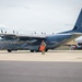 A military C-130 Hercules transport aircraft with tail code 'LI' and serial number 870 is parked on a flight line apron. A single service member in military fatigues is walking in front of the aircraft carrying bright orange wheel chocks or safety cones. Emergency/fire response vehicles are visible in the background near a hangar facility.