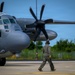 A military ground crew member in flight suit and hearing protection walks toward a large gray turboprop military transport aircraft on a tarmac. The aircraft, marked with the number '5870', appears to be a C-130 Hercules variant, with its large six-blade propeller prominently visible. The scene takes place on an airfield apron with green vegetation visible in the background under a clear blue sky.