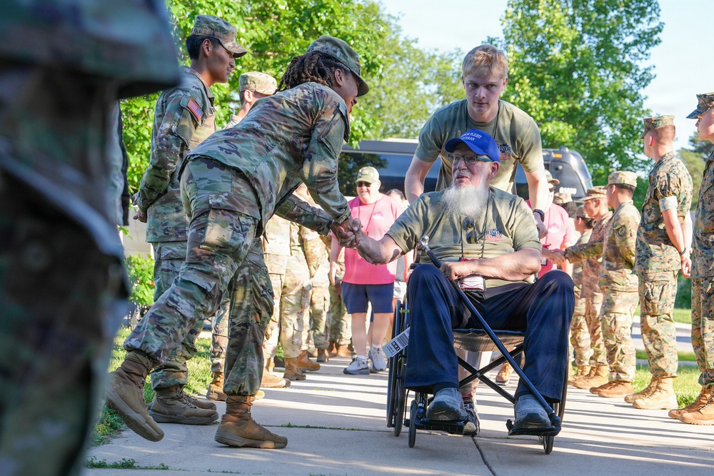 A veteran in a wheelchair is being greeted by a line of military personnel in camouflage uniforms on a paved walkway. A soldier is leaning forward to shake or fist-bump the elderly bearded man's hand in a show of respect. A young civilian male is pushing the wheelchair while onlookers observe in the background.
