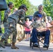 A veteran in a wheelchair is being greeted by a line of military personnel in camouflage uniforms on a paved walkway. A soldier is leaning forward to shake or fist-bump the elderly bearded man's hand in a show of respect. A young civilian male is pushing the wheelchair while onlookers observe in the background.