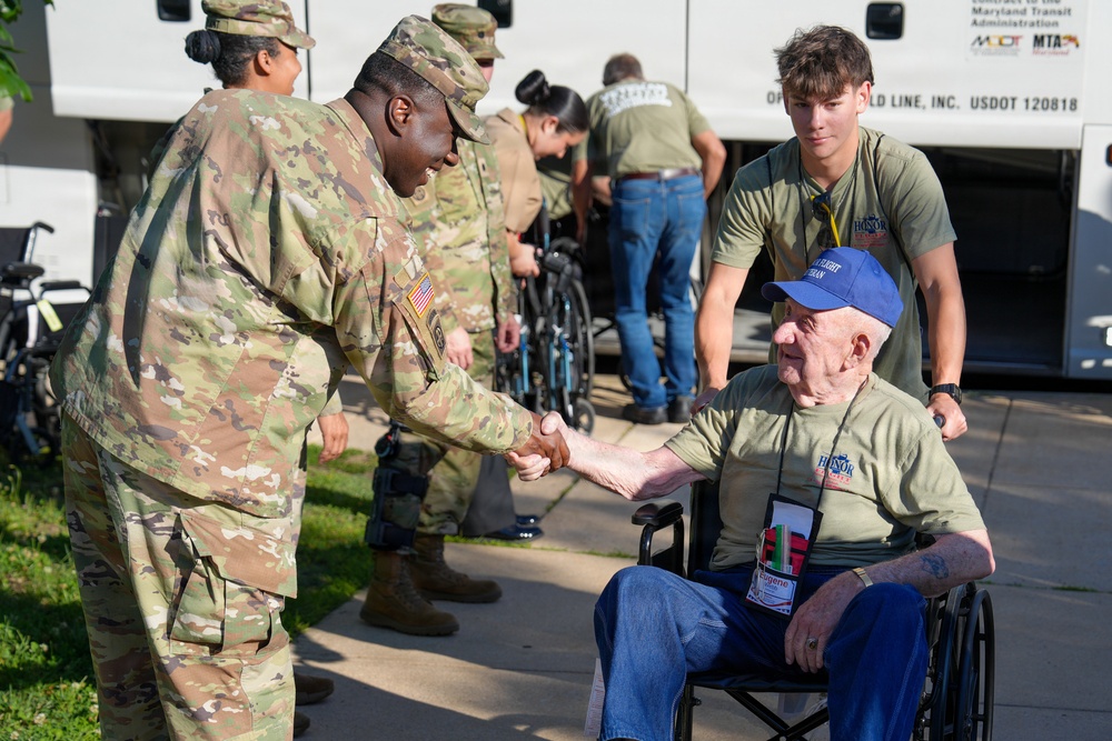 A U.S. Army soldier in OCP (Operational Camouflage Pattern) uniform is shaking hands with an elderly veteran seated in a wheelchair, who is wearing a blue cap and an olive green 'Honor Flight' t-shirt with a name badge reading 'Eugene'. A young civilian volunteer in a matching Honor Flight shirt assists the veteran from behind, while additional military personnel and wheelchairs are visible near a bus in the background.