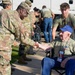 A U.S. Army soldier in OCP (Operational Camouflage Pattern) uniform is shaking hands with an elderly veteran seated in a wheelchair, who is wearing a blue cap and an olive green 'Honor Flight' t-shirt with a name badge reading 'Eugene'. A young civilian volunteer in a matching Honor Flight shirt assists the veteran from behind, while additional military personnel and wheelchairs are visible near a bus in the background.