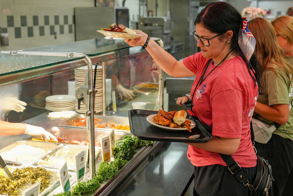 A woman in a red t-shirt and glasses is serving herself food at a cafeteria-style buffet line. She is holding a tray with a plate of what appears to be chicken and a roll, while reaching for an additional plate above the sneeze guard. A food service worker wearing white gloves is visible behind the counter.