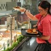 A woman in a red t-shirt and glasses is serving herself food at a cafeteria-style buffet line. She is holding a tray with a plate of what appears to be chicken and a roll, while reaching for an additional plate above the sneeze guard. A food service worker wearing white gloves is visible behind the counter.