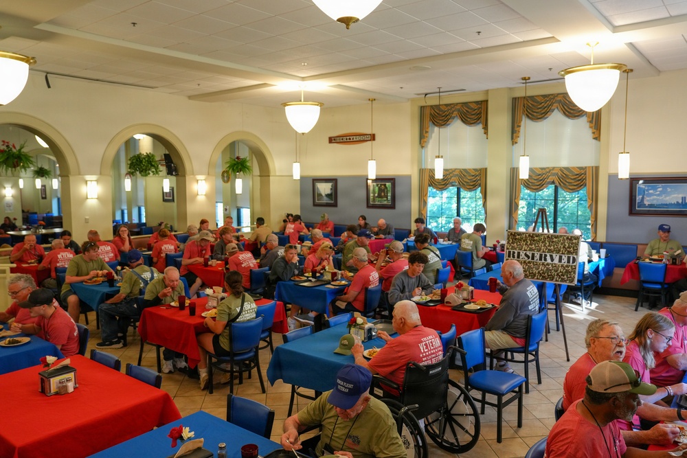 A large dining hall filled with a group of veterans and their companions sharing a meal together. The majority of attendees are wearing matching red or olive/gray t-shirts that appear to read 'VETERAN' on the back, suggesting an organized honor flight or veterans' recognition event. The room features blue and red tablecloths, arched architectural details, and a 'RESERVED' sign near the windows.