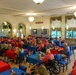 A large dining hall filled with a group of veterans and their companions sharing a meal together. The majority of attendees are wearing matching red or olive/gray t-shirts that appear to read 'VETERAN' on the back, suggesting an organized honor flight or veterans' recognition event. The room features blue and red tablecloths, arched architectural details, and a 'RESERVED' sign near the windows.