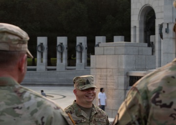 Three U.S. Army soldiers in MultiCam/OCP camouflage uniforms are engaged in conversation at the National World War II Memorial in Washington, D.C. The central soldier is facing the camera and smiling, while the other two soldiers are seen from behind. The setting appears to be during evening hours based on the lighting conditions.