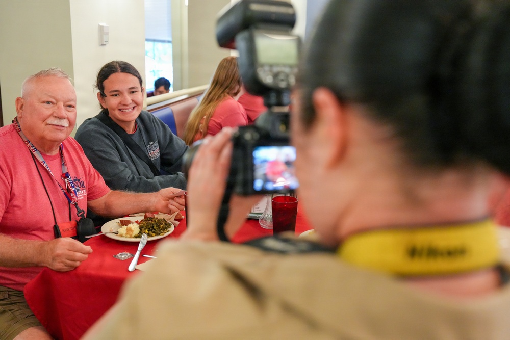 A photographer with a Nikon camera is photographing two people seated at a table with a red tablecloth in what appears to be a dining hall or restaurant. An older man in a pink shirt and a younger woman in a gray sweatshirt are posing together and smiling for the photo. The scene suggests a casual social or institutional dining event.