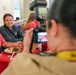 A photographer with a Nikon camera is photographing two people seated at a table with a red tablecloth in what appears to be a dining hall or restaurant. An older man in a pink shirt and a younger woman in a gray sweatshirt are posing together and smiling for the photo. The scene suggests a casual social or institutional dining event.
