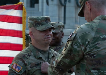 A military ceremony is taking place outdoors, likely an award or promotion ceremony. A senior officer is pinning something onto the uniform of a soldier identified by the name tape 'LIVELY', with an American flag and a third soldier visible in the background. The setting appears to be a formal military installation or memorial grounds.