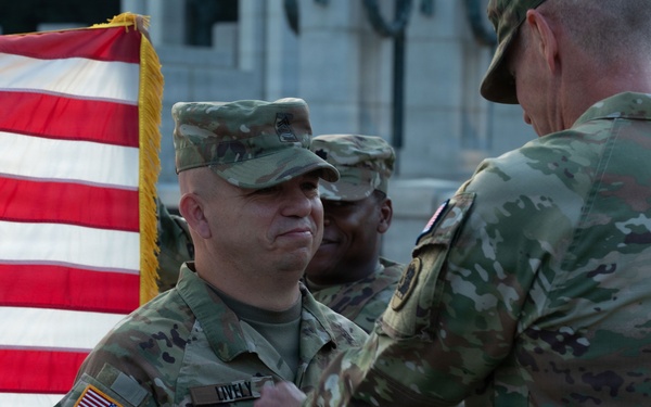 A military ceremony is taking place outdoors, likely an award or promotion ceremony. A senior officer is pinning something onto the uniform of a soldier identified by the name tape 'LIVELY', with an American flag and a third soldier visible in the background. The setting appears to be a formal military installation or memorial grounds.