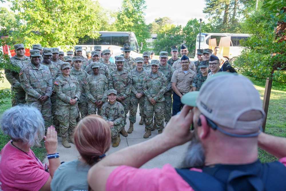 A large group of military personnel in various uniforms, primarily Army camouflage (OCP pattern) with some Navy and Air Force members, pose for a group photograph outdoors. Three civilians in the foreground — two women and a man — are photographing or observing the group. The setting appears to be a military installation or outdoor facility with buses and trees visible in the background.