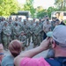 A large group of military personnel in various uniforms, primarily Army camouflage (OCP pattern) with some Navy and Air Force members, pose for a group photograph outdoors. Three civilians in the foreground — two women and a man — are photographing or observing the group. The setting appears to be a military installation or outdoor facility with buses and trees visible in the background.