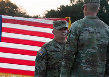 Two U.S. Army soldiers in OCP (Operational Camouflage Pattern) uniforms stand facing each other in front of a large American flag with gold fringe. The setting appears to be the National World War II Memorial in Washington, D.C., based on the visible stone architecture in the background. The scene is captured at sunset or golden hour, with civilians visible in the background.