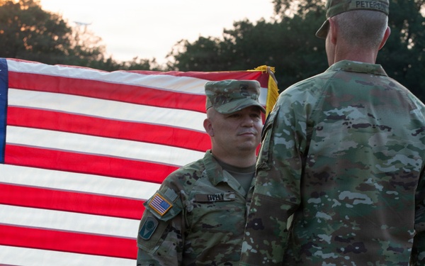 Two U.S. Army soldiers in OCP (Operational Camouflage Pattern) uniforms stand facing each other in front of a large American flag with gold fringe. The setting appears to be the National World War II Memorial in Washington, D.C., based on the visible stone architecture in the background. The scene is captured at sunset or golden hour, with civilians visible in the background.