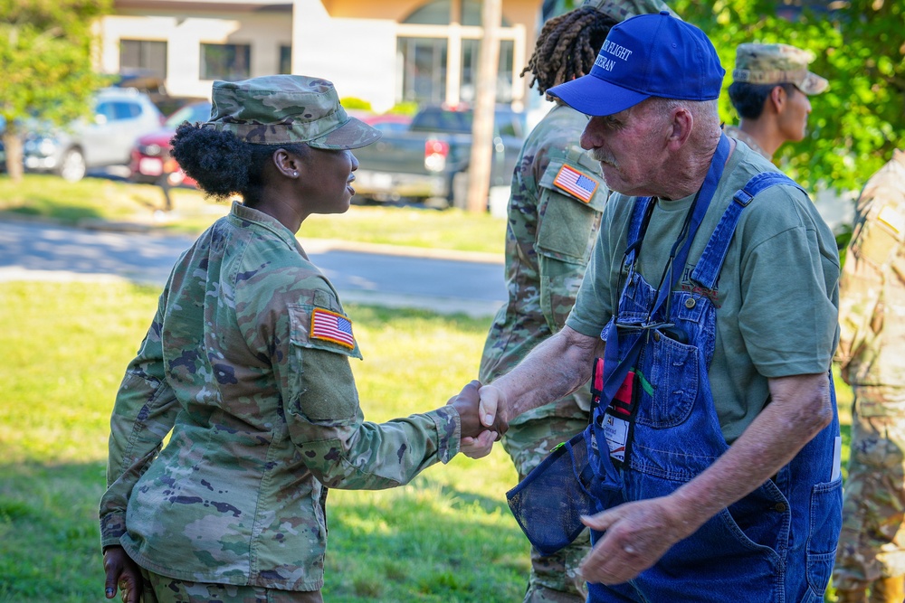 A U.S. Army soldier in OCP (Operational Camouflage Pattern) uniform is shaking hands with an older civilian man wearing denim overalls and a blue baseball cap. The interaction appears cordial and respectful, taking place outdoors on a grassy area near a parking lot. Additional soldiers in uniform are visible in the background.
