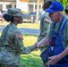 A U.S. Army soldier in OCP (Operational Camouflage Pattern) uniform is shaking hands with an older civilian man wearing denim overalls and a blue baseball cap. The interaction appears cordial and respectful, taking place outdoors on a grassy area near a parking lot. Additional soldiers in uniform are visible in the background.