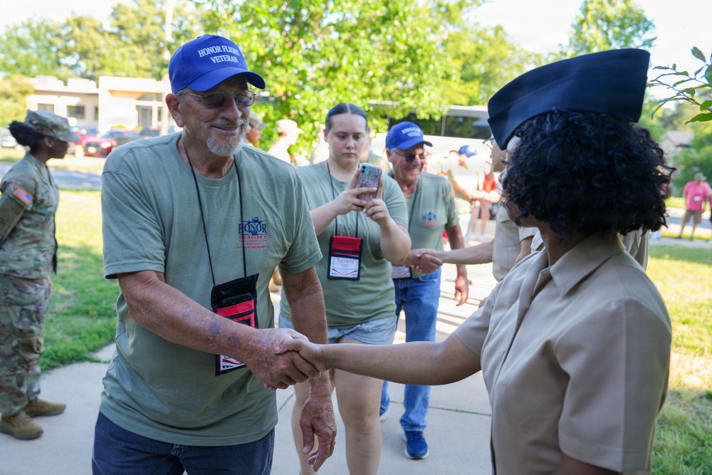 A group of Honor Flight veterans wearing blue caps and olive green shirts with lanyards are being greeted by a uniformed military or ROTC member in tan uniform and dark garrison cap. The primary focus is a handshake between an older male veteran and the uniformed individual. A woman in the background is photographing the event on a smartphone, and a U.S. Army soldier in camouflage uniform is visible on the left.