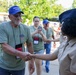 A group of Honor Flight veterans wearing blue caps and olive green shirts with lanyards are being greeted by a uniformed military or ROTC member in tan uniform and dark garrison cap. The primary focus is a handshake between an older male veteran and the uniformed individual. A woman in the background is photographing the event on a smartphone, and a U.S. Army soldier in camouflage uniform is visible on the left.