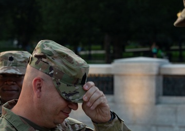 Three U.S. Army soldiers in MultiCam/OCP camouflage uniforms are gathered outdoors, likely during a military ceremony or promotion event. The central soldier, identified by name tape as 'LIVELY,' is adjusting or receiving his patrol cap, suggesting a pinning or promotion ceremony. The setting appears to be a formal outdoor military event near a monument or memorial.