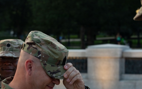 Three U.S. Army soldiers in MultiCam/OCP camouflage uniforms are gathered outdoors, likely during a military ceremony or promotion event. The central soldier, identified by name tape as 'LIVELY,' is adjusting or receiving his patrol cap, suggesting a pinning or promotion ceremony. The setting appears to be a formal outdoor military event near a monument or memorial.