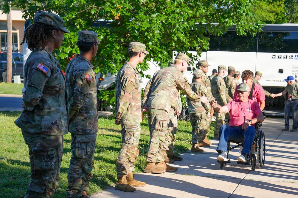 A line of U.S. Army soldiers in OCP (Operational Camouflage Pattern) uniforms are greeting a civilian man seated in a wheelchair, who is wearing a pink shirt and green cap. The soldiers appear to be shaking hands with the individual as he moves along the formation. The setting appears to be an outdoor area near a building, with a white bus visible in the background.