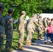 A line of U.S. Army soldiers in OCP (Operational Camouflage Pattern) uniforms are greeting a civilian man seated in a wheelchair, who is wearing a pink shirt and green cap. The soldiers appear to be shaking hands with the individual as he moves along the formation. The setting appears to be an outdoor area near a building, with a white bus visible in the background.