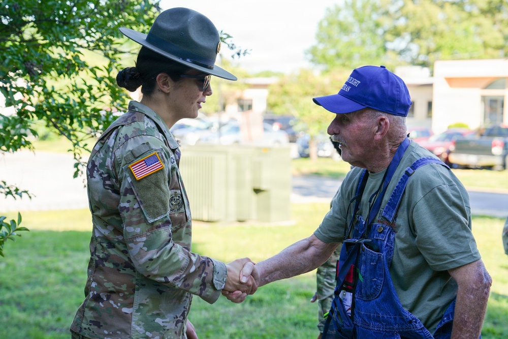 A female U.S. Army soldier in full camouflage uniform and drill sergeant hat is shaking hands with an older civilian man wearing blue overalls and a blue baseball cap. The interaction appears cordial and takes place outdoors on a grassy area near a parking lot. The soldier displays a U.S. flag patch on her right sleeve, consistent with active U.S. Army dress standards.