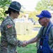 A female U.S. Army soldier in full camouflage uniform and drill sergeant hat is shaking hands with an older civilian man wearing blue overalls and a blue baseball cap. The interaction appears cordial and takes place outdoors on a grassy area near a parking lot. The soldier displays a U.S. flag patch on her right sleeve, consistent with active U.S. Army dress standards.