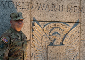 A U.S. Army soldier in MultiCam (OCP) camouflage uniform stands in front of the World War II Memorial in Washington, D.C. The memorial's granite wall features a carved Great Seal eagle relief with the Latin motto 'E Pluribus Unum.' The soldier is posing for a photograph beside the iconic memorial entrance marker.