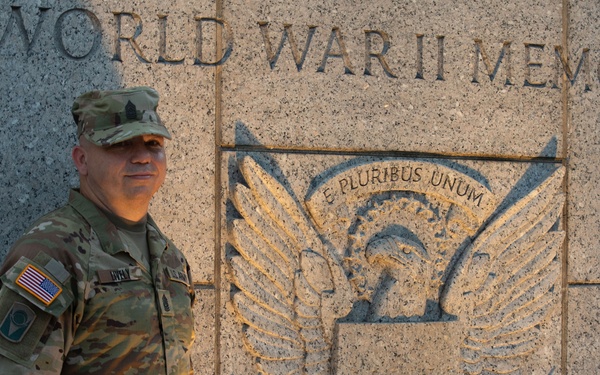 A U.S. Army soldier in MultiCam (OCP) camouflage uniform stands in front of the World War II Memorial in Washington, D.C. The memorial's granite wall features a carved Great Seal eagle relief with the Latin motto 'E Pluribus Unum.' The soldier is posing for a photograph beside the iconic memorial entrance marker.