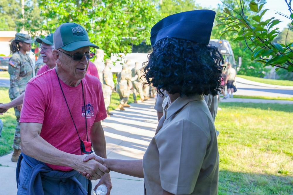An elderly civilian man wearing a pink 'Honor Flight' t-shirt and green cap is shaking hands with a military service member in a tan dress uniform and navy blue garrison cap. In the background, a group of soldiers in camouflage uniforms are lined up along a sidewalk, suggesting a formal welcome or greeting ceremony. The setting appears to be an outdoor military or civic event on a sunny day.