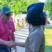 An elderly civilian man wearing a pink 'Honor Flight' t-shirt and green cap is shaking hands with a military service member in a tan dress uniform and navy blue garrison cap. In the background, a group of soldiers in camouflage uniforms are lined up along a sidewalk, suggesting a formal welcome or greeting ceremony. The setting appears to be an outdoor military or civic event on a sunny day.