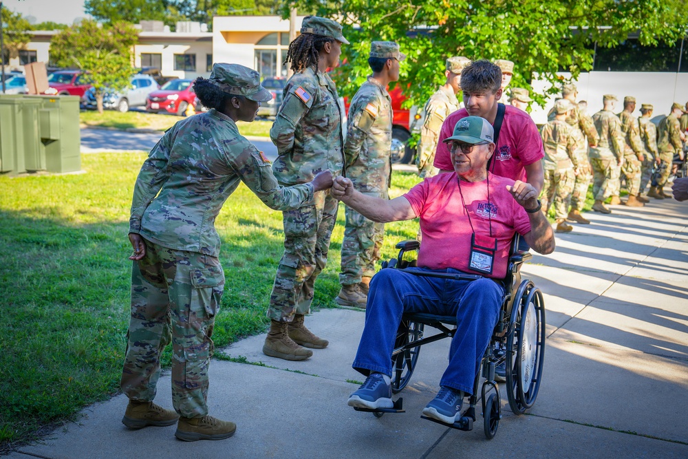 A U.S. Army soldier in OCP (Operational Camouflage Pattern) uniform is giving a fist bump to an elderly civilian man seated in a wheelchair, who is wearing a pink shirt and a lanyard with a badge. The civilian appears to be a veteran participating in what looks like an Honor Flight or similar veteran recognition program. Multiple soldiers in uniform are visible in the background forming a line, suggesting a formal greeting or welcoming ceremony.
