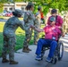 A U.S. Army soldier in OCP (Operational Camouflage Pattern) uniform is giving a fist bump to an elderly civilian man seated in a wheelchair, who is wearing a pink shirt and a lanyard with a badge. The civilian appears to be a veteran participating in what looks like an Honor Flight or similar veteran recognition program. Multiple soldiers in uniform are visible in the background forming a line, suggesting a formal greeting or welcoming ceremony.
