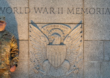 A U.S. Army soldier in MultiCam (OCP) camouflage uniform stands at attention beside the World War II Memorial wall in Washington, D.C. The granite wall features a large carved Great Seal of the United States with the motto 'E Pluribus Unum' and the inscription 'WORLD WAR II MEMORIAL' above it. The image appears to be an official or commemorative photograph taken at the national monument.